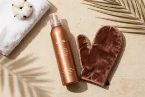 Self-tanner mousse bottle and velvet tanning mitt on travertine tile with sunlit palm shadows